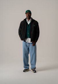 Svart fleecejacka med gröna accenter, grön väst över vit skjorta, ljusblå lösa jeans och svarta sneakers. Grön keps komplettar looken.