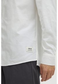 Camisa blanca de manga larga hecha de algodón suave. Presenta una abertura lateral y una etiqueta en el dobladillo inferior. Tejido ligeramente texturizado. Pantalones oscuros visibles.