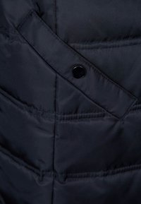 Marinblå pufferjacka i ett tyg med kviltad textur, med ett diagonalt lock och svarta tryckknappar. Slätt, hållbart material.