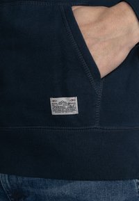 Marineblauwe sweatshirt met een kangoeroezak, ribgebreide boorden en zoom. Bevat een wit merklabel aan de onderkant aan de linkerkant. Zachte textuur.