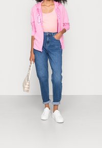 Veste à capuche zippée rose avec logo, débardeur rose clair, jean bleu à revers, et baskets blanches ; tenant un sac à main beige à rayures.