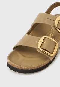 Μπεζ σουέντ σανδάλι με δύο φαρδιές λουράκια, μεγάλες χρυσές αγκράφες, διαμορφωμένη φελλό σόλα και μαύρη υφασμάτινη σόλα.