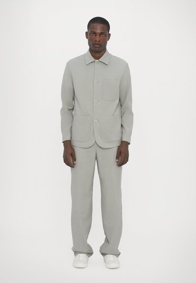 Lichtgrijze, op maat gemaakte blazer met een kraag, knopen aan de voorkant en een borstzak, gecombineerd met bijpassende broeken. Gemaakt met witte schoenen.