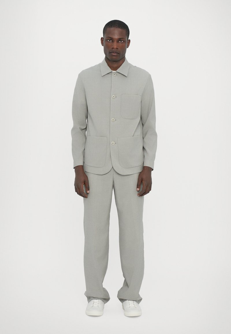 Ανοιχτό γκρι προσαρμοσμένο σακάκι με κολάρο, μπροστινά κουμπιά και τσέπη στο στήθος, συνδυασμένο με αντίστοιχες παντελόνες. Φοριέται με λευκά παπούτσια.