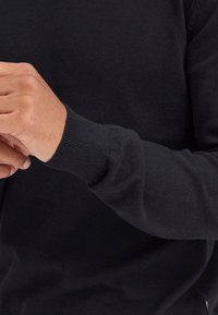 Czarny sweter z dzianiny, zbliżenie na mankiet i dłoń. Miękka tekstura i prążkowany detal przy nadgarstku. Prosty, nieskomplikowany design.