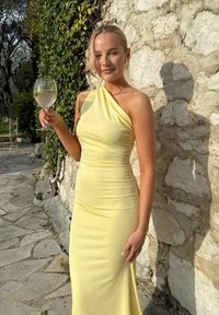 Žena v žltých šatách na jedno rameno drží pohár s plátkom limetky, stojac pri kamennej stene, na ktorú dopadá slnečné svetlo a vrhá jej tieň.