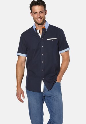 Kurzärmliges, marinefarbenes Hemd mit Knöpfen, hellblauen Akzenten, weißer Brusttasche und strukturiertem Kragen. Kombiniert mit blauen Jeans.