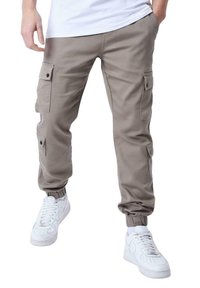 Pantalones cargo de color marrón claro con puños elásticos, que cuentan con múltiples bolsillos laterales y botones a presión, combinados con zapatillas blancas.