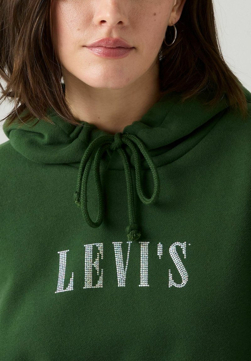 Grön fleecehoodie med dragsko i huvan. Har silverfärgade rhinestones i bokstäverna "LEVI'S" på framsidan. Slät textur, avslappnad design.