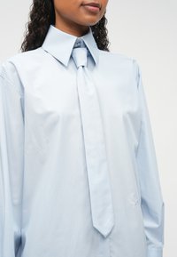 Ανοιχτό μπλε πουκάμισο με μεγάλο γιακά και αντίστοιχο γραβάτα. Φτιαγμένο από απαλό ύφασμα, διαθέτει κλείσιμο με κουμπιά και διακριτική κεντημένη λογότυπο.