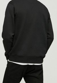 Sudadera negra con un cuello redondo y puños de canalé, con un acabado texturizado. Llevada sobre una camisa blanca y combinada con pantalones negros.