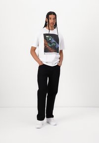 Tricou alb cu o grafică colorată a unui reptil, asociat cu pantaloni negri și adidași albi. Modelul stă cu mâinile în buzunare.