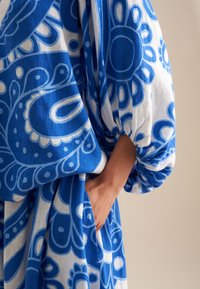 Abito blu e bianco con motivo floreale, dotato di maniche larghe, tessuto arricciato ai polsi e una tasca laterale. Materiale leggero e textured.
