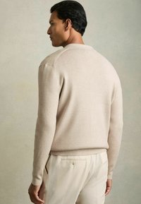 Beige ribbad tröja med långa ärmar, figurnära design. Slät textur. Buren med ljusa byxor, sedd bakifrån.