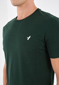 Tamnozeleni pamučni majica s okruglim izrezom i kratkim rukavima. Sadrži mali bijeli vez ptice na lijevoj strani prsa. Glatka tekstura.
