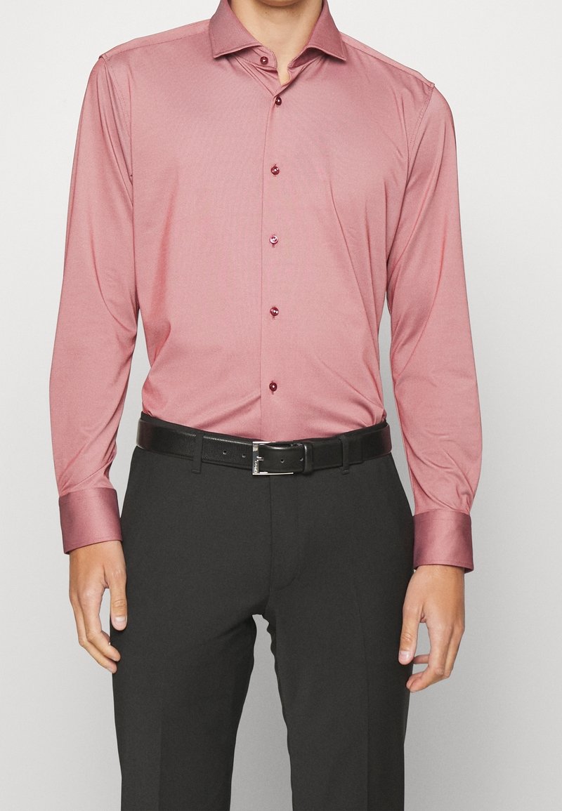 Uomo che indossa una camicia rosa a maniche lunghe, aderente, infilata in pantaloni neri con una cintura di pelle nera.