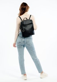 Svart läderryggsäck med texturerad yta, rektangulär form, justerbara remmar och en framficka med dragkedja, vilande på en ljus denimoutfit.