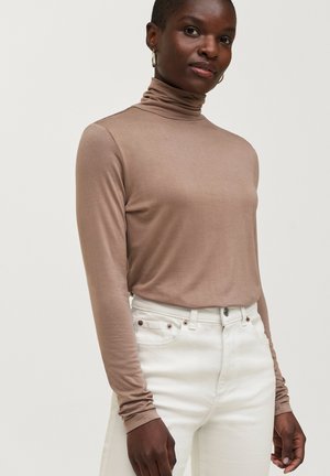 Langærmet turtleneck-top i lys brun, fremstillet af et glat stof, parret med tætsiddende hvide jeans med metalknapdetaljer.