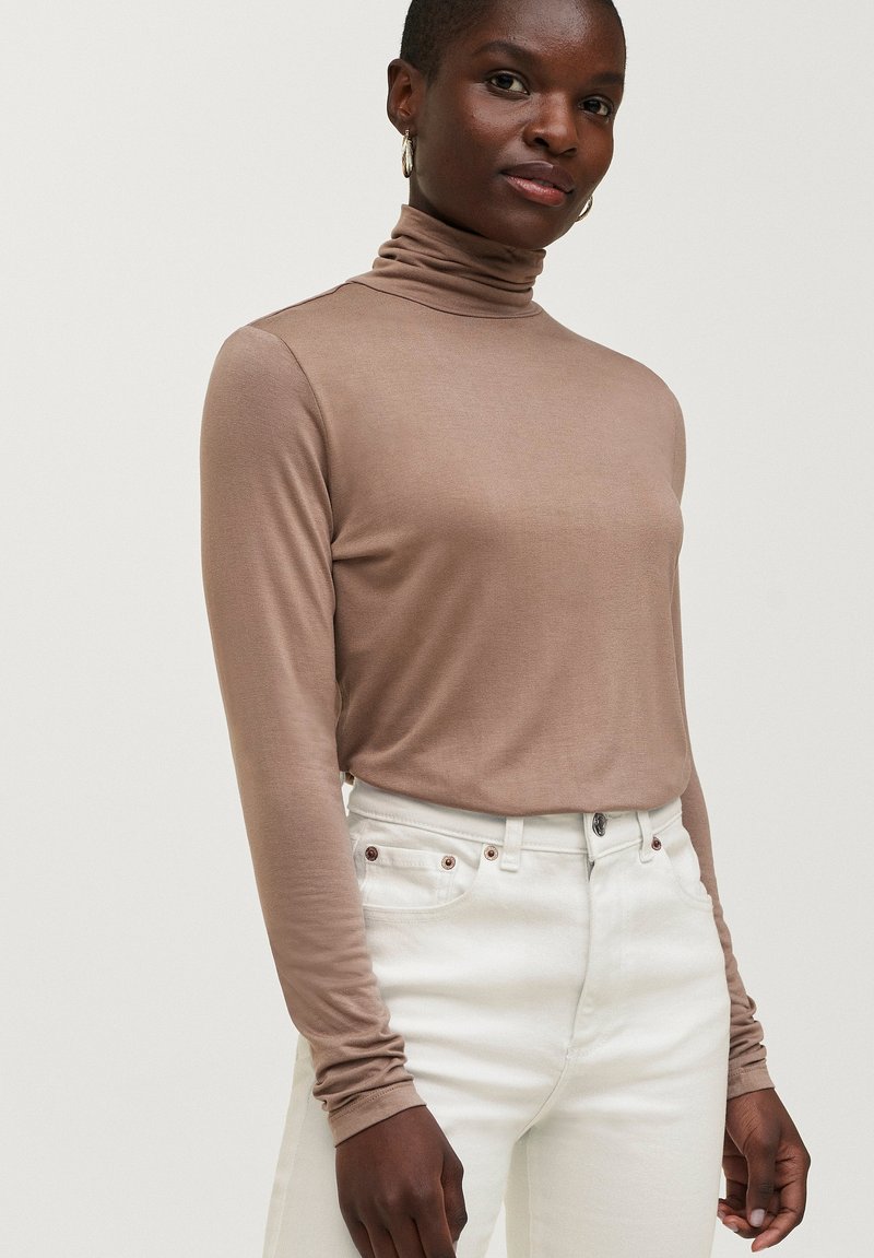 Langærmet turtleneck-top i lys brun, fremstillet af et glat stof, parret med tætsiddende hvide jeans med metalknapdetaljer.