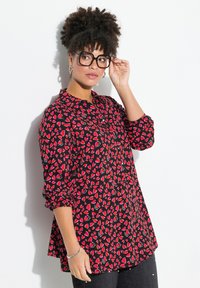 Rot und schwarz kariertes Hemd mit Herzmuster, langen Ärmeln und lockerer Passform. Das Model trägt schwarze Brille und silberne Accessoires.