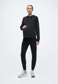 Czarna bluza z kapturem o luźnym kroju, zestawiona z dopasowanymi czarnymi spodniami. Model nosi jasnoszare buty sportowe, prezentując wygodny, casualowy look.