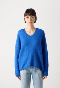 Blue knitted sweater with a V-neck, loose fit, and ribbed texture. Features long sleeves and a small metallic detail at the hem.