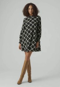 Melnbalts rakstains kleita ar garām piedurknēm, augstu kaklu un brīvu piegriezumu, apvienota ar ceļgaliem līdz anklei skaidriem zābakiem.