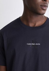 Navy blue cotton t-shirt with a round neckline. Features a printed logo "Calvin Klein Jeans" and an accentuated "CK" in reflective detail.