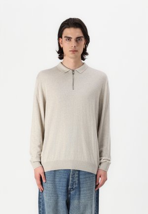 Beige long-sleeve polo shirt with a zippered collar, ribbed cuffs, and hem. Worn over loose, faded blue jeans. Smooth texture, light fabric.