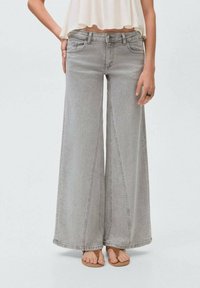 Breda jeans i ljusgrå denim med en blekt finish. Har framfickor och dekorativ sömnad längs benen. Platta sandaler bärs.
