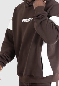 Personne portant un sweat à capuche marron avec des rayures blanches sur les manches et le texte "SMILODOX" sur la poitrine, ajustant la capuche près de son visage.