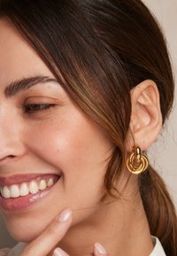 Goldene Creolen mit ineinander verlinkten, kreisförmigen Designs in polierter Optik, vor einem neutralen Hintergrund.