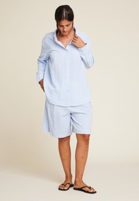 Lyseblå skjorte med lange ermer, kombinert med matchende løse shorts. Glatt stoff, avslappet passform og svarte flate sandaler.