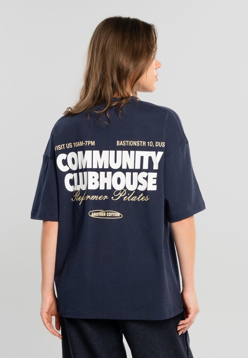 Ung kvinde med brunt hår iført en navyblå oversized t-shirt med hvid og beige tekst, der promoverer et Community Clubhouse Pilates-studie.
