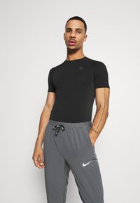 Schwarzes Sportshirt mit kurzen Ärmeln und strukturiertem Stoff, kombiniert mit grauen Hosen, die ein weißes Nike-Logo zeigen. Das Modell steht mit Händen in den Taschen.
