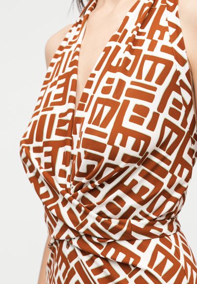 Γυναίκα που φορά αμάνικο φόρεμα με βαθύ V-λαιμόκοψη, με καφέ και λευκό γεωμετρικό σχέδιο και ρινόμενη ύφανση στη μέση.