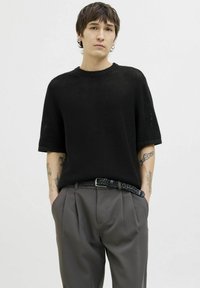 Black knit short-sleeve sweater with a loose fit, paired with tailored gray trousers and a braided black belt; visible wrist tattoos.