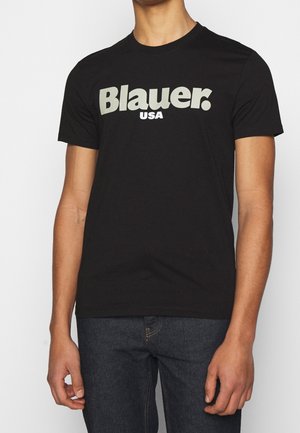 Person trägt ein schwarzes T-Shirt mit kurzen Ärmeln und dem Schriftzug "Blauer. USA" in großen weißen Buchstaben, kombiniert mit dunkelblauen Jeans.