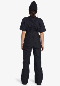 Osoba v černé čepici, černém tričku s krátkým rukávem a černých sněhových kalhotách s kříženými kšandami, stojící zády k bílé stěně.