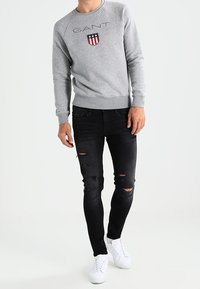Sudadera gris con logo "GANT" y gráfico de escudo; combinada con vaqueros ajustados negros rasgados y zapatillas blancas. Estilo deportivo casual.