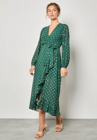 Abito avvolgente verde a pois con scollatura a V profonda, maniche lunghe a sbuffo, orlo arricciato e dettaglio in vita annodato, realizzato in un tessuto leggero.