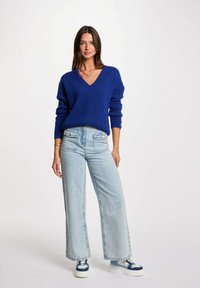 Maglione blu a trama con scollo a V, abbinato a jeans a gamba larga di un blu chiaro e sneakers blu navy e bianche. Accento con una semplice collana.