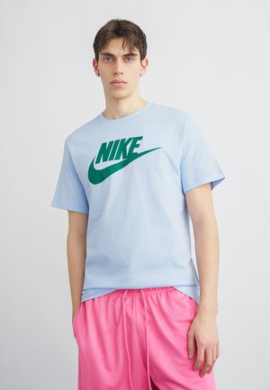 Jeune homme portant un t-shirt Nike bleu clair et un short rose vif, debout devant un fond clair uni.