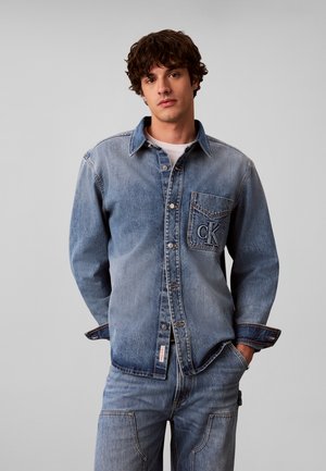 Junger Mann mit lockigem Haar, der ein ausgewaschenes blaues Denimhemd mit "CK"-Logo auf der Brusttasche und passende Denim-Jeans trägt, Hände in den Taschen.