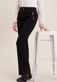 Pantalons noirs taille haute avec un top ajusté et des jambes évasées. Dotés de boutons métalliques sur le côté et d'un tissu lisse et texturé.