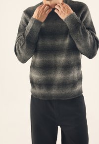 Pull en maille grise avec des rayures horizontales subtiles, col rond et poignets côtelés. Associé à un pantalon noir, mettant en valeur une finition texturée.