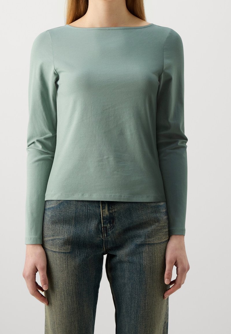 Personne portant une chemise vert clair à manches longues et un jean bleu délavé, debout, les bras détendus le long du corps sur un fond blanc uni.