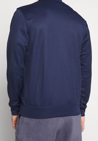 Hombre con un sudadera de mangas largas azul marino y pantalones grises, visto desde la espalda contra un fondo claro y liso.