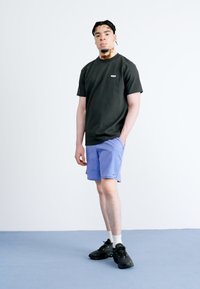 Black short-sleeve t-shirt with logo, paired with light purple shorts and black sneakers. Model stands on a blue floor against a neutral background.