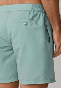 Bañador corto para hombre de color verde claro con cintura elástica, bolsillo lateral y bolsillo trasero con botón, usado por una persona de pie.